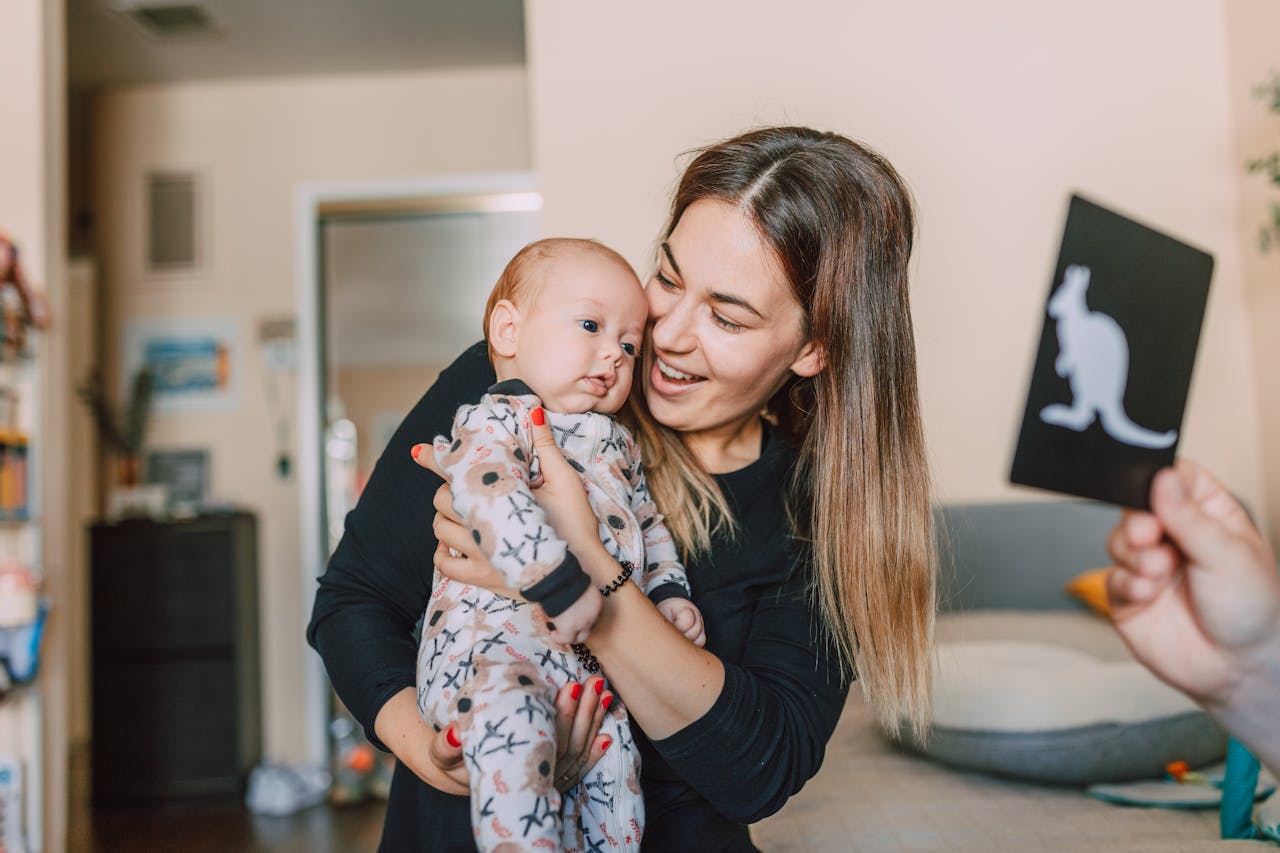 A joyful mother holds her baby, engaging them with a kangaroo flashcard indoors.