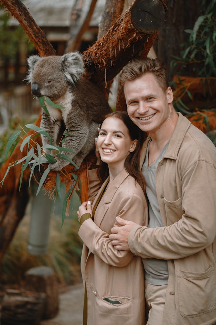 Side view of cheerful stylish couple embracing each other and looking at camera while standing near funny cute koala in koala sanctuary in Australia